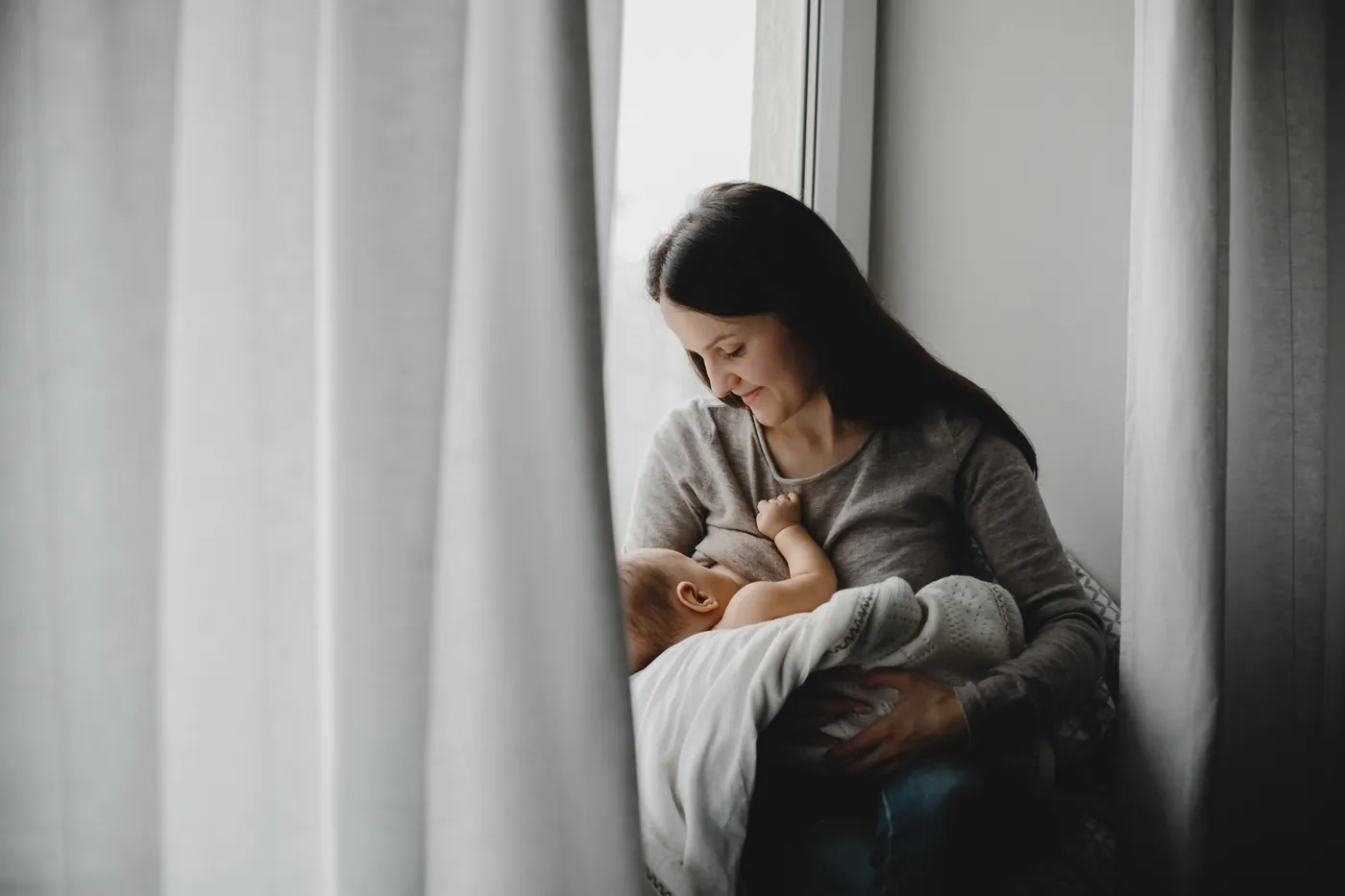 Une mère allaite son nouveau-né, assise près d'une fenêtre.