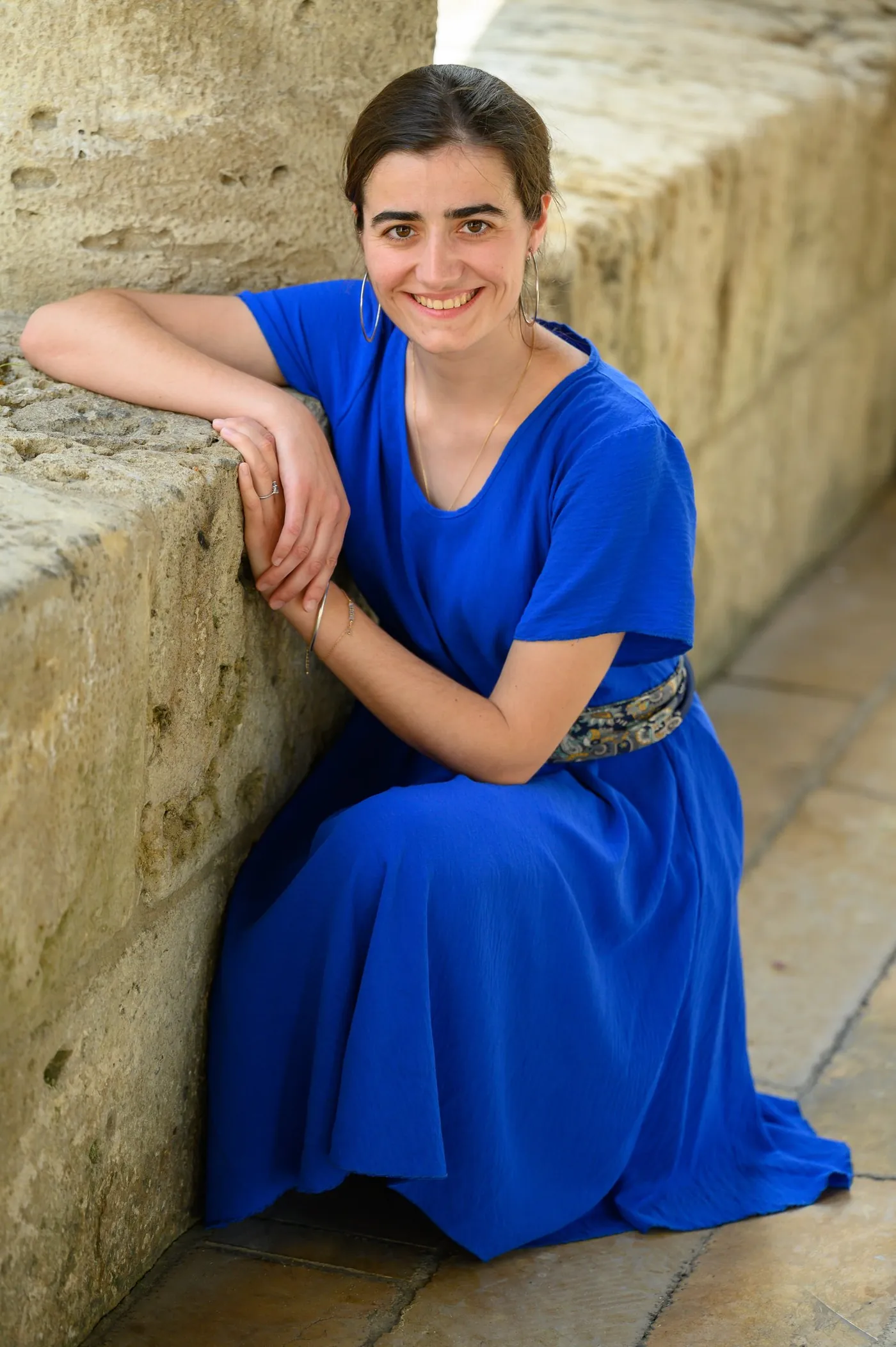 Quiterie de Butler, souriante, en robe bleue, assise contre un mur de pierre.